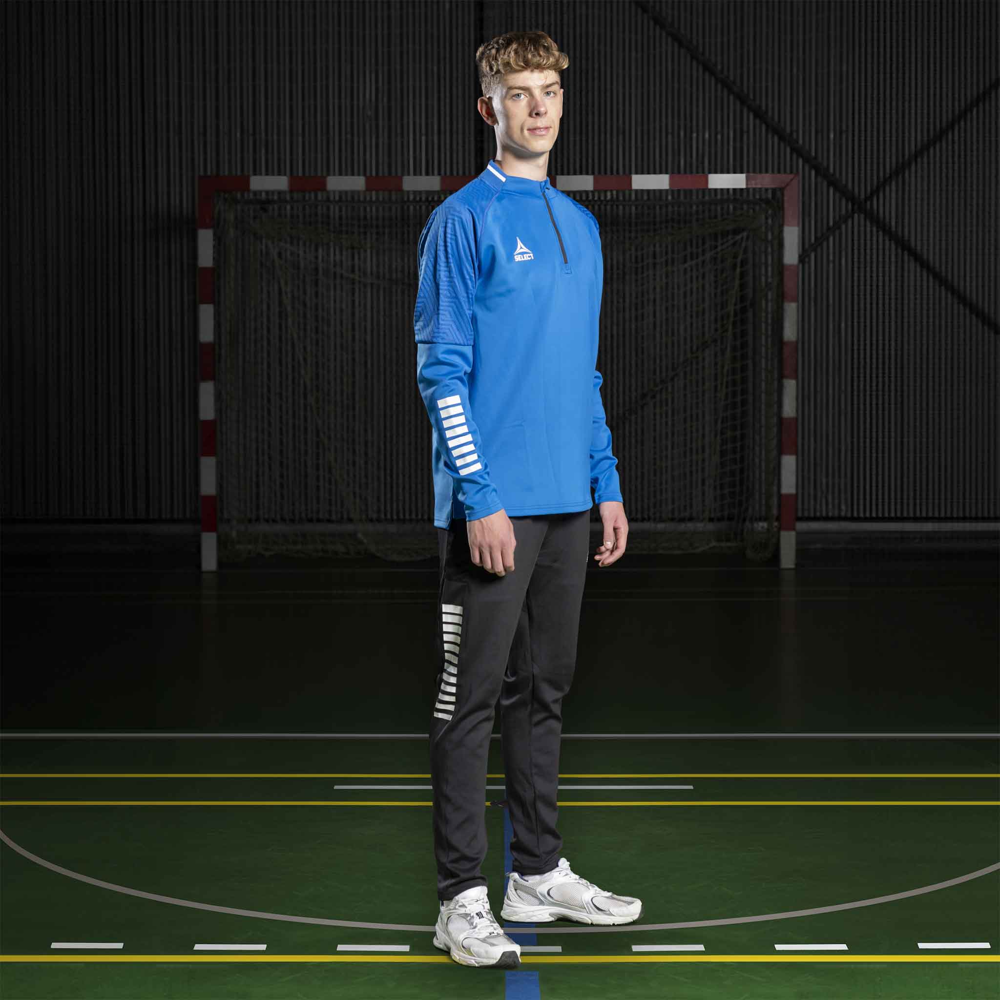 Manlig handbollsspelare i blå Training sweat 1/2 zip Monaco v24 och svarta byxor, Match & Training wear, framifrån.