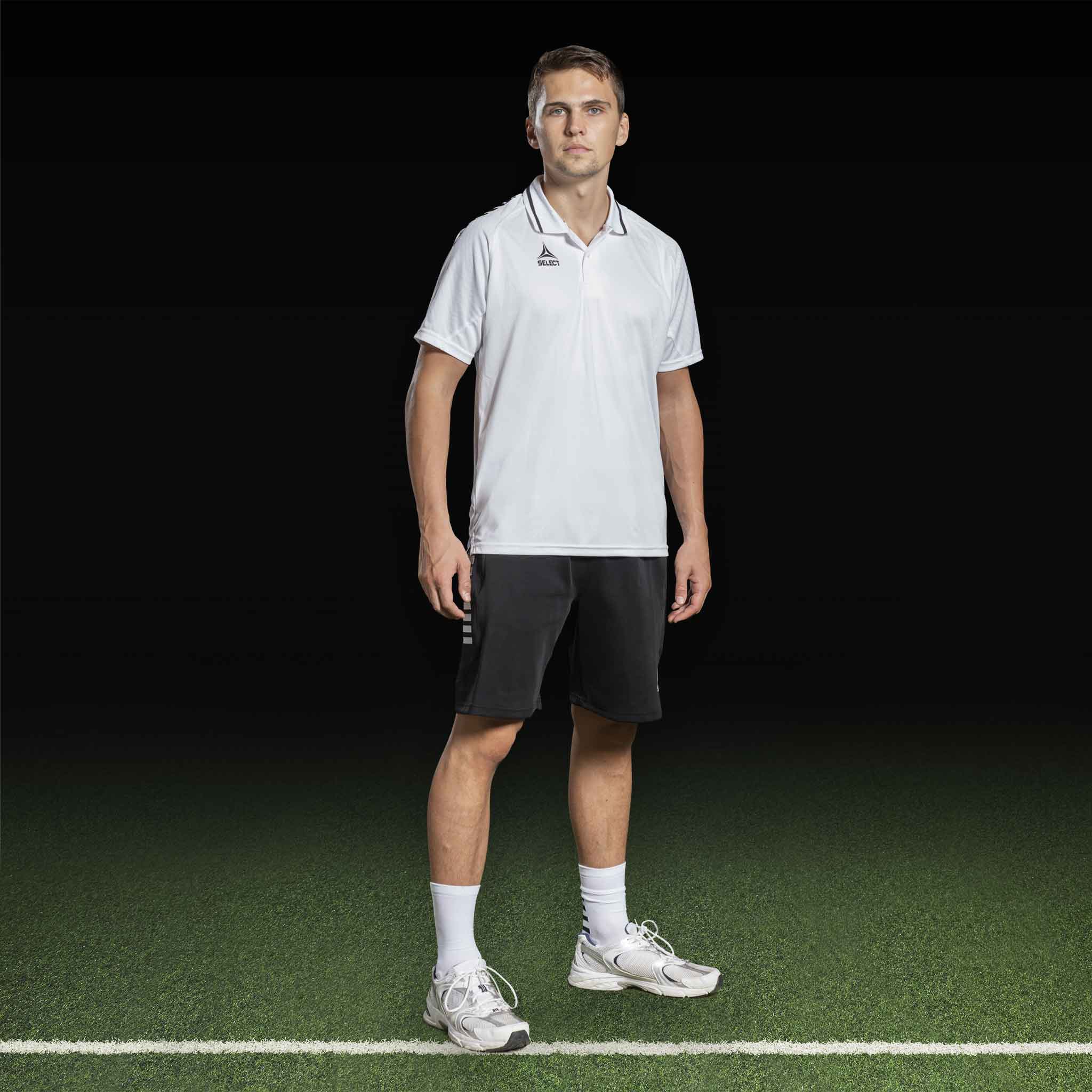 Manlig fotbollsspelare i vit piké och svarta Bermuda shorts Monaco v24 fritidskläder på plan.