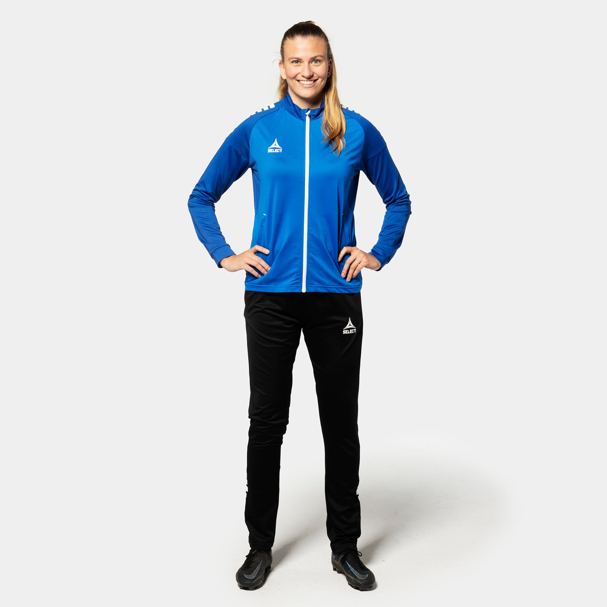 Kvinnlig fotbollsspelare i blåvit Training zip jacket Spain woman v25, Match & Training-kläder, med svarta byxor, sedd framifrån.