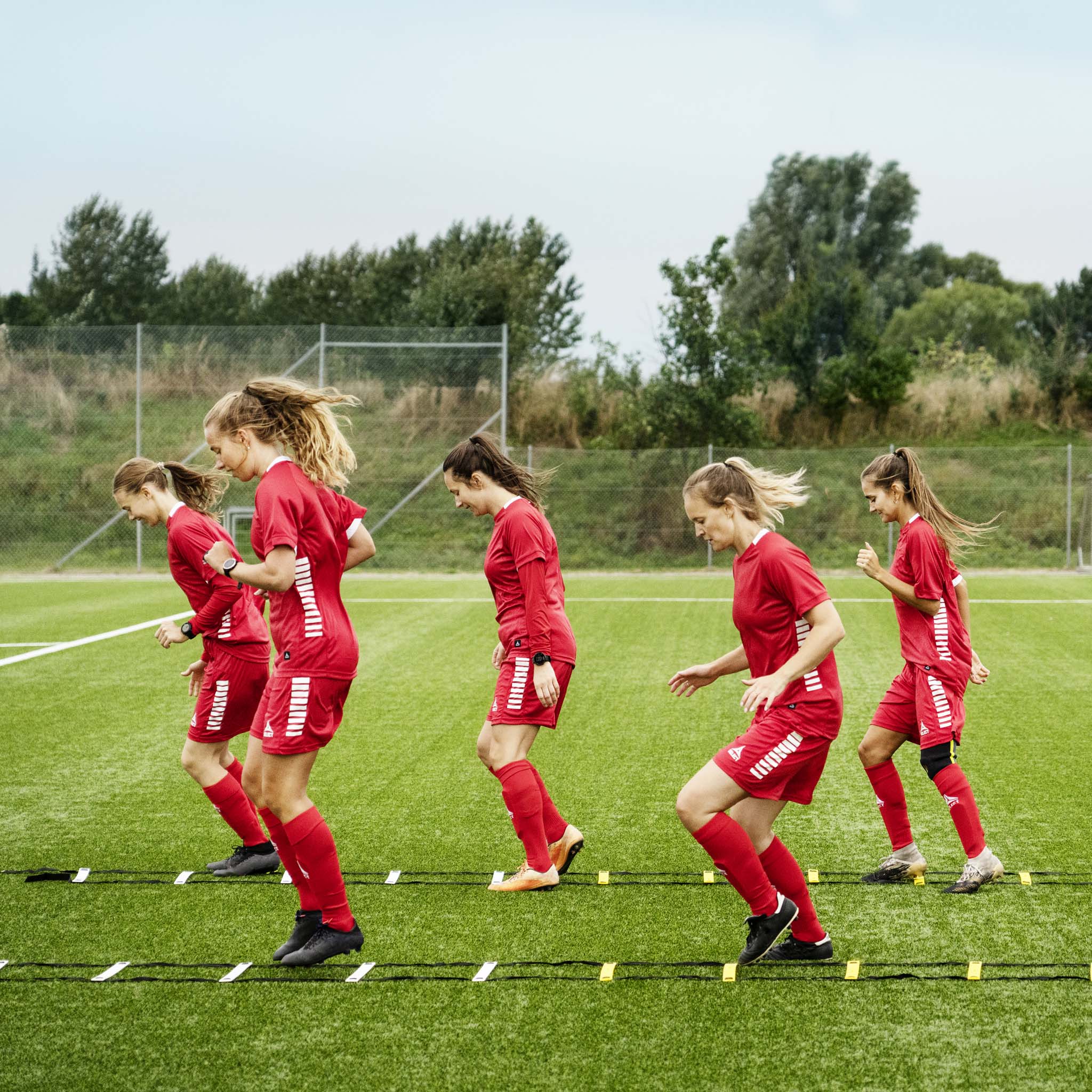 Kvinnliga fotbollsspelare tränar med orange och gul Agility ladder, fotbollsträningsutrustning, på grön plan.